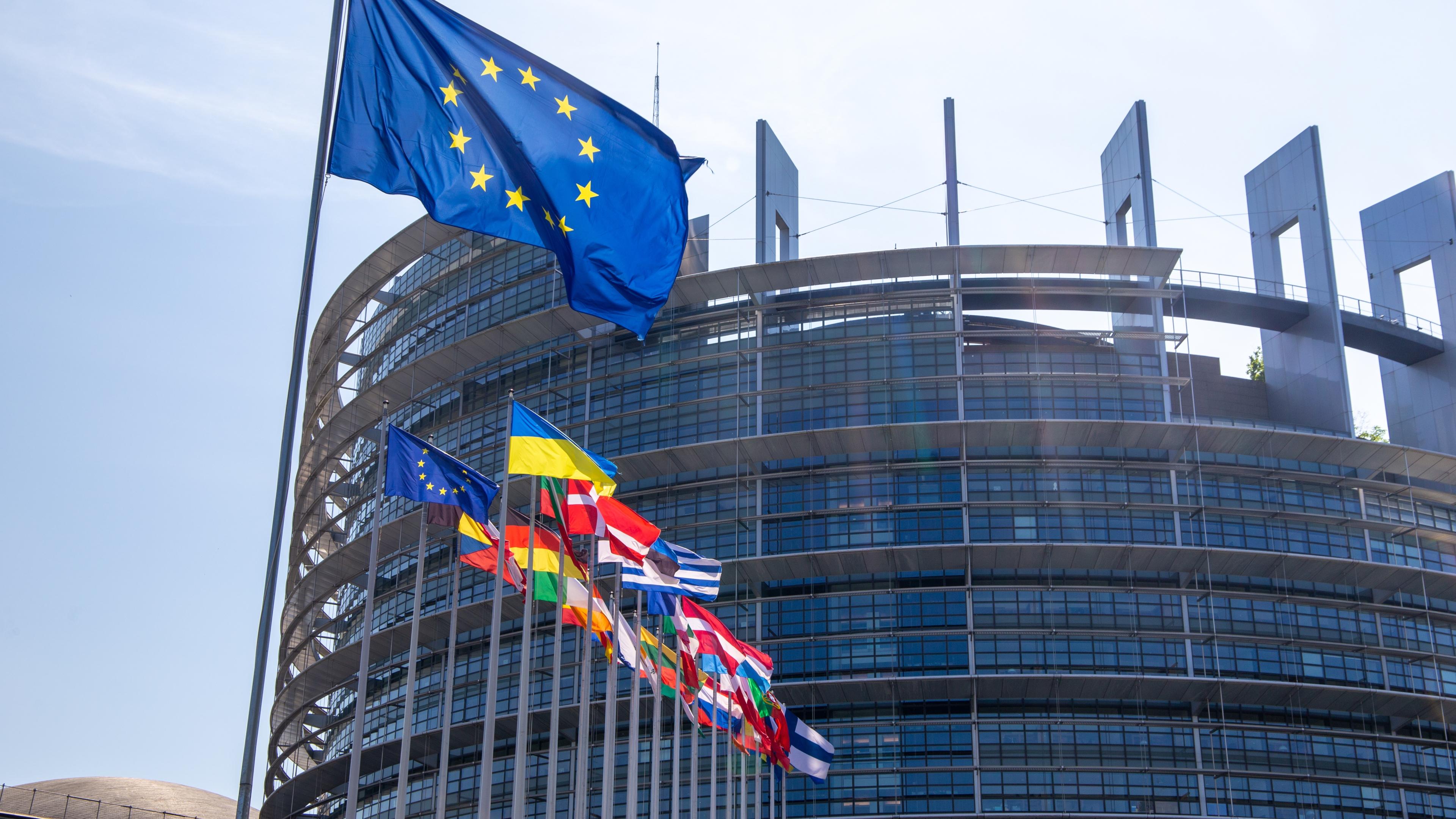 Das europäische Parlament bei Tageslicht und mit der europäischen Flagge im Vordergrund.